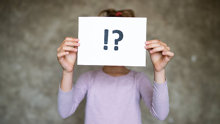 FAQ Ein Kind hält ein Blatt mit einem großen Fragezeichen und einem großen Ausrufezeichen vor sein Gesicht